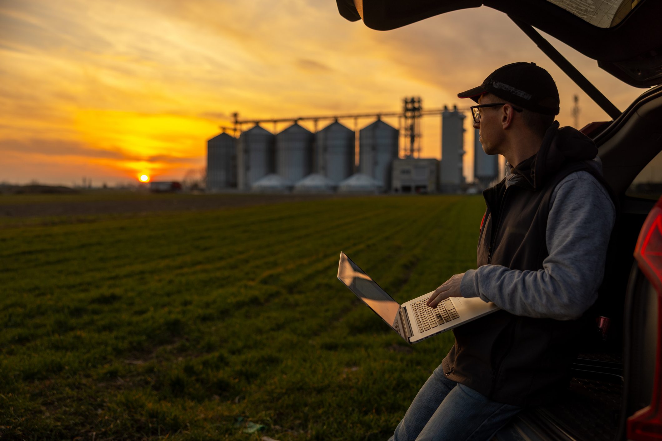 Caucasian Farmer Working on Laptop in Field at Sunset Working on Agriculture Equipment